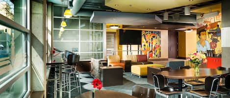 A modern hotel lobby with a mix of seating areas, including a round table with chairs, a sofa, and a lounge with a colorful mural.