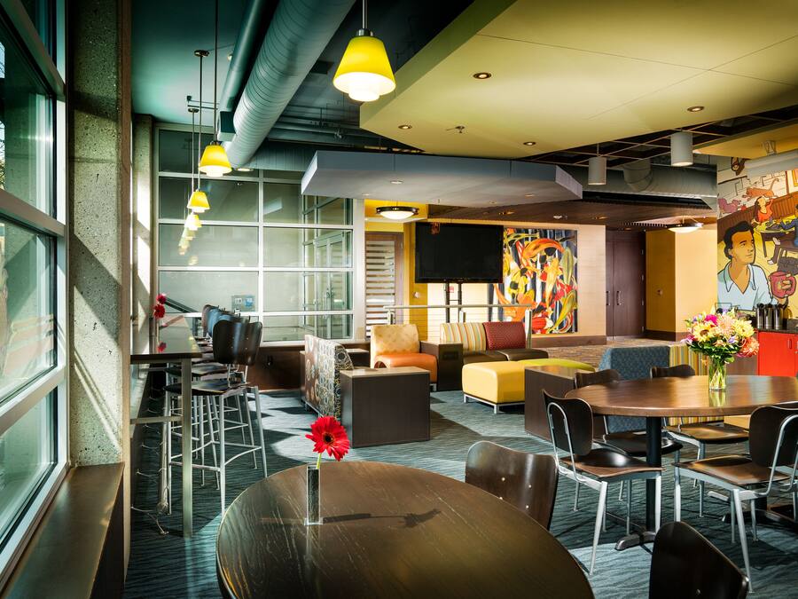 Un moderno vestíbulo de hotel con diversas zonas de estar, incluyendo una mesa redonda con sillas, un sofá y un área de descanso con un mural colorido.
