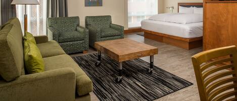 Habitación de hotel con cama, sofá, dos sillones, una mesa de centro de madera, lámpara y televisor.