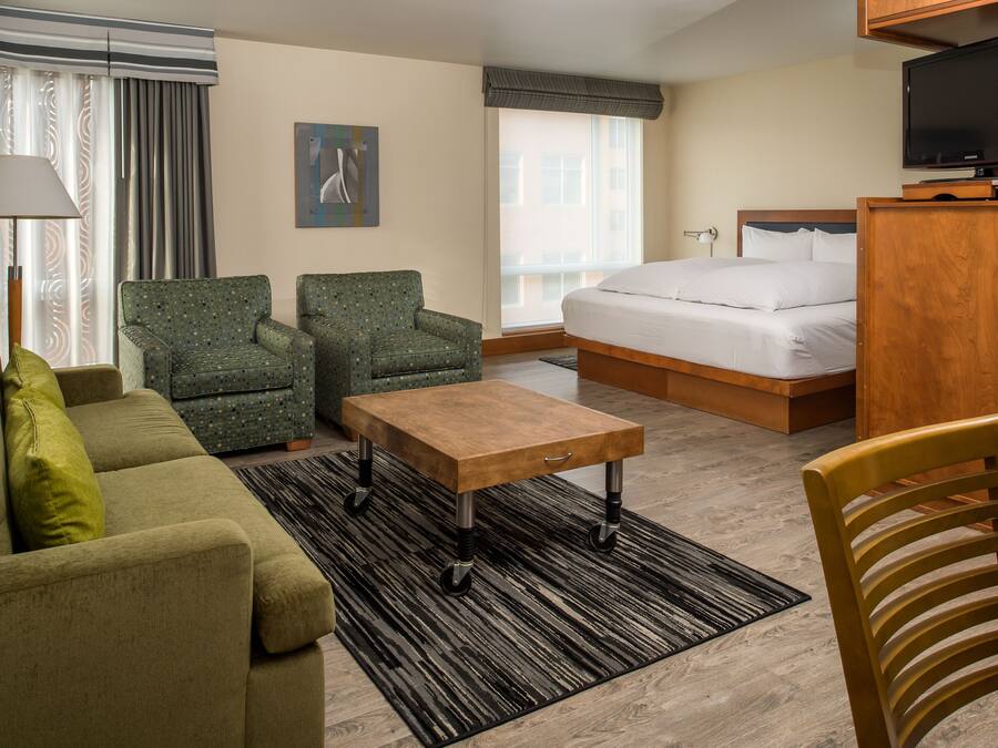 Habitación de hotel con cama, sofá, dos sillones, una mesa de centro de madera, lámpara y televisor.