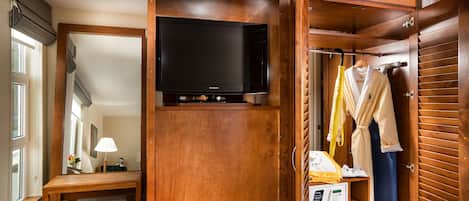 A hotel room with a wooden wardrobe, a television, a bench, a lamp, and a clothing rack with robes and shoes.