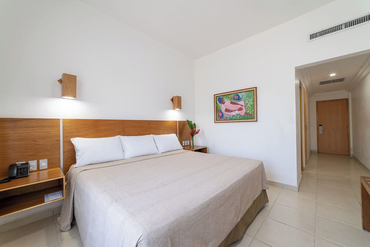 A hotel room with a large bed, wooden headboard, bedside table with a phone, a small nightstand with a lamp, a painting on the wall, and a door leading to another room.