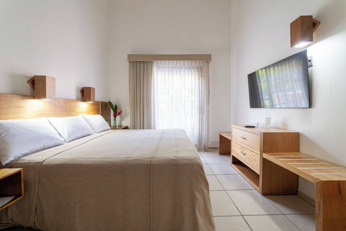 A hotel room with a large bed, wooden headboard, a flat-screen TV mounted on the wall, a wooden bench, and a desk with drawers.