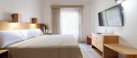 A hotel room with a large bed, wooden headboard, a flat-screen TV mounted on the wall, a wooden bench, and a desk with drawers.