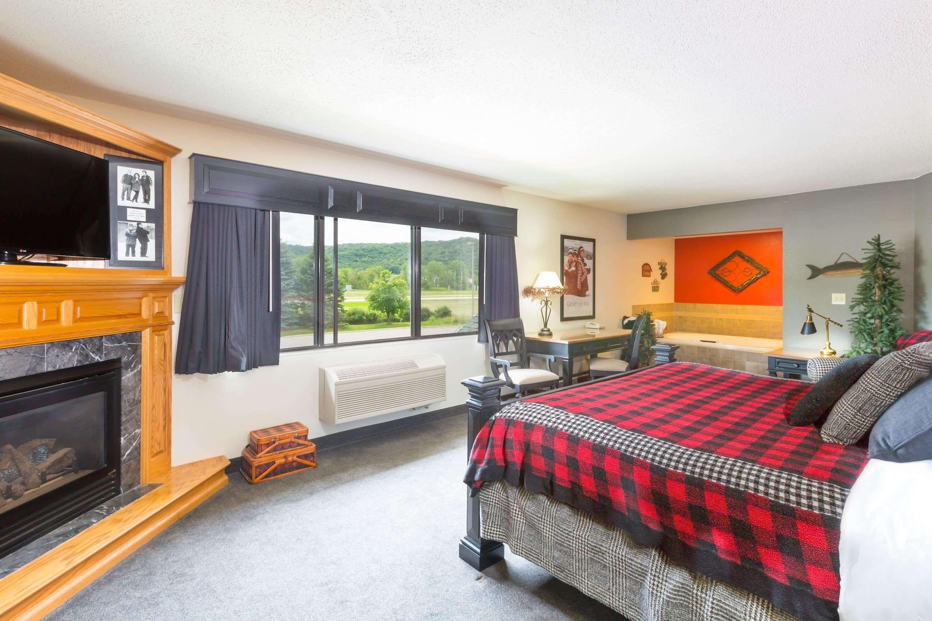 Habitación de hotel con chimenea, una cama con manta a cuadros, un escritorio con lámpara y vistas al exterior.