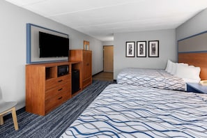 A hotel room with a TV, wooden wardrobe, two beds with patterned bedding, a small desk, and framed pictures on the wall.