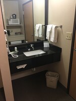 Un baño de hotel con un lavabo negro, un espejo, un tendedero de toallas con toallas blancas, un cesto de basura y un secador de pelo en un estante.