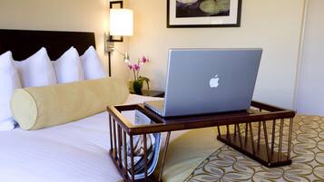 Habitación de hotel con cama, una laptop sobre una bandeja, una lámpara y un cuadro en la pared.