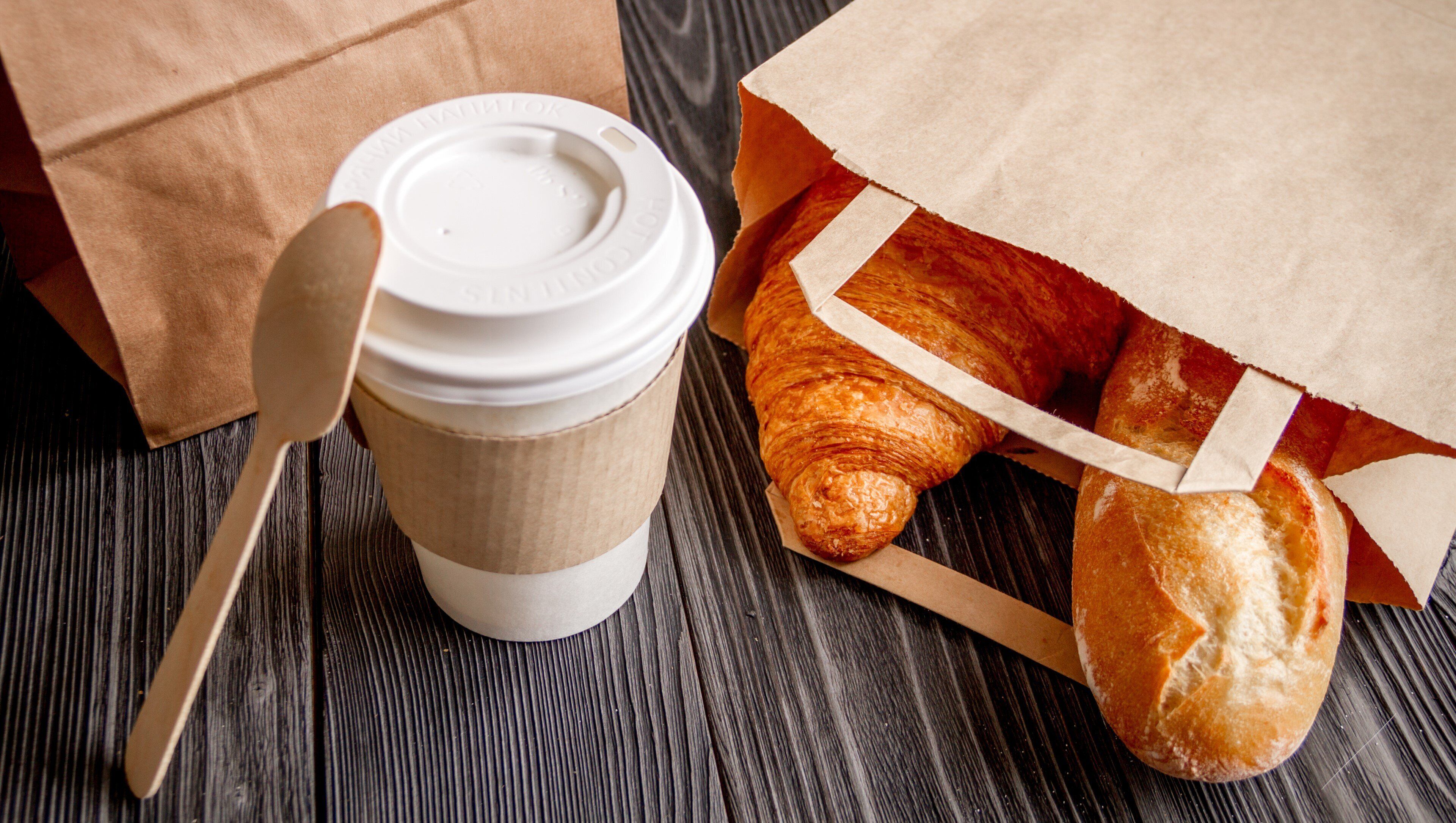 Una taza de café de papel con tapa y una cuchara, junto a una bolsa de papel y un croissant.