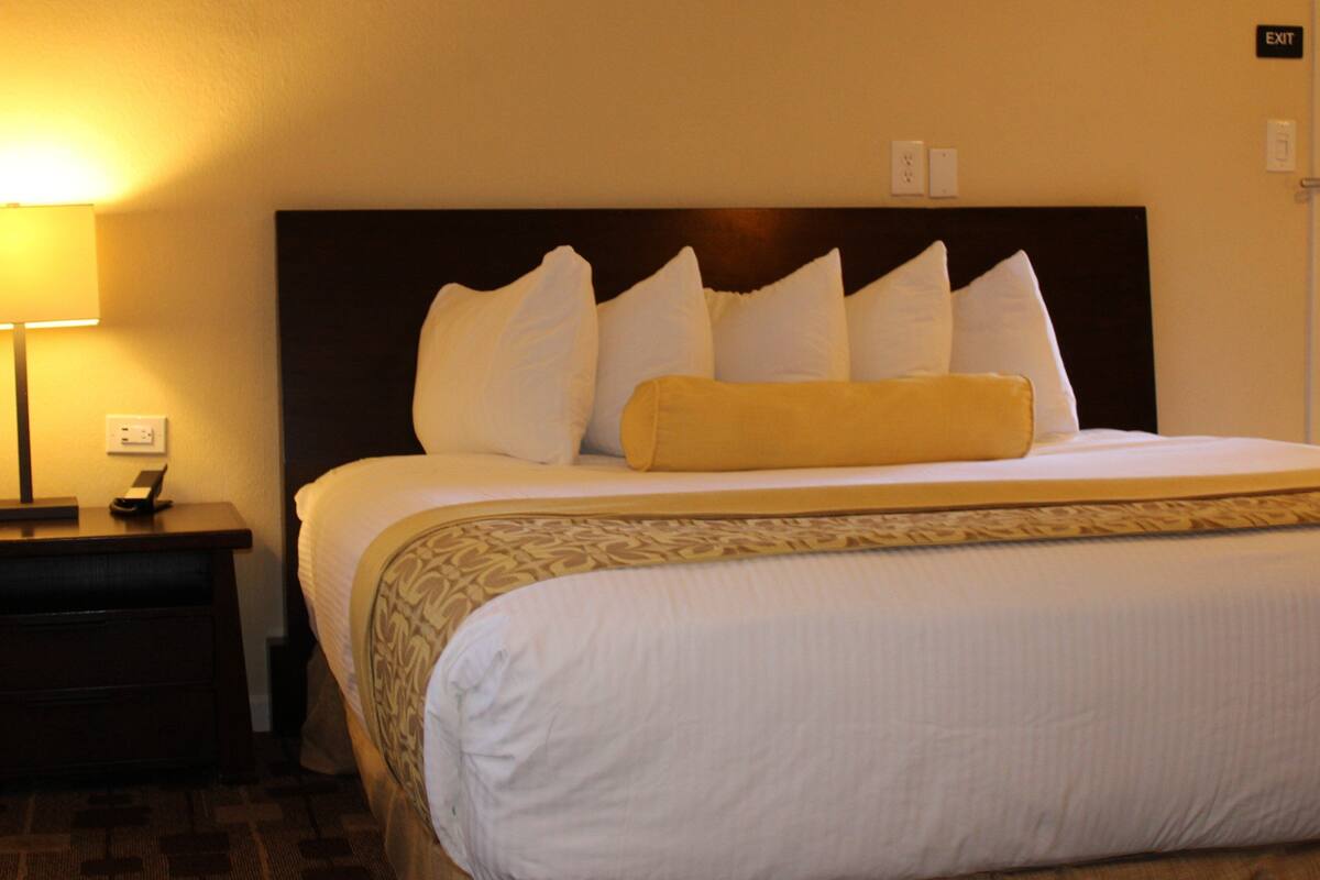 A hotel room with a neatly made bed, a bedside lamp, a clock, and an exit sign on the wall.