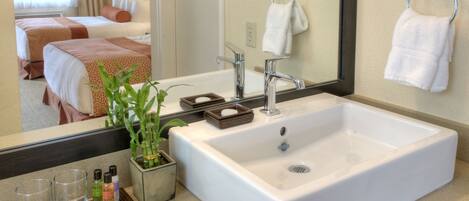 A hotel bathroom with a large sink, a mirror, a towel rack, and a bed in the background.