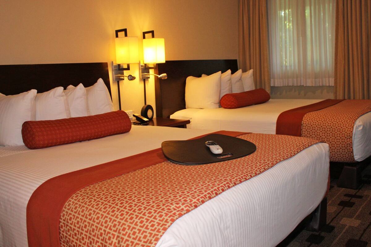 A hotel room with two beds, each with white linens and red accents, a bedside table with a remote control, and a window with curtains.