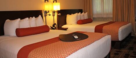 A hotel room with two beds, each with white linens and red accents, a bedside table with a remote control, and a window with curtains.
