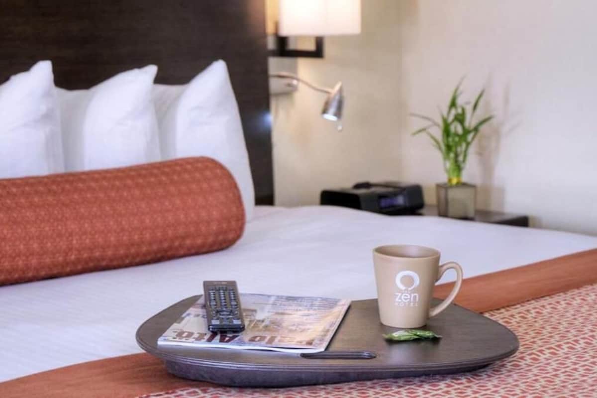 Una cama bien hecha con almohadas blancas y rojas, un control remoto, una revista y una taza en una bandeja.