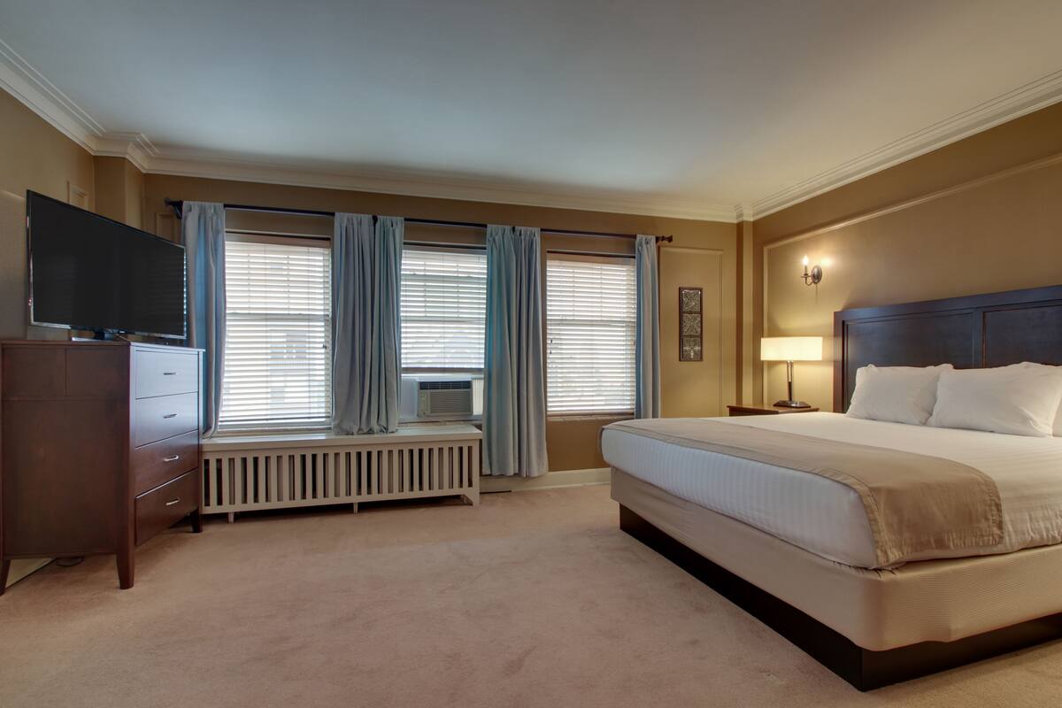 Ein Hotelzimmer mit einem großen Bett, einem Schminktisch, einem Fernseher, einem Fenster mit Vorhängen und einer Heizung.