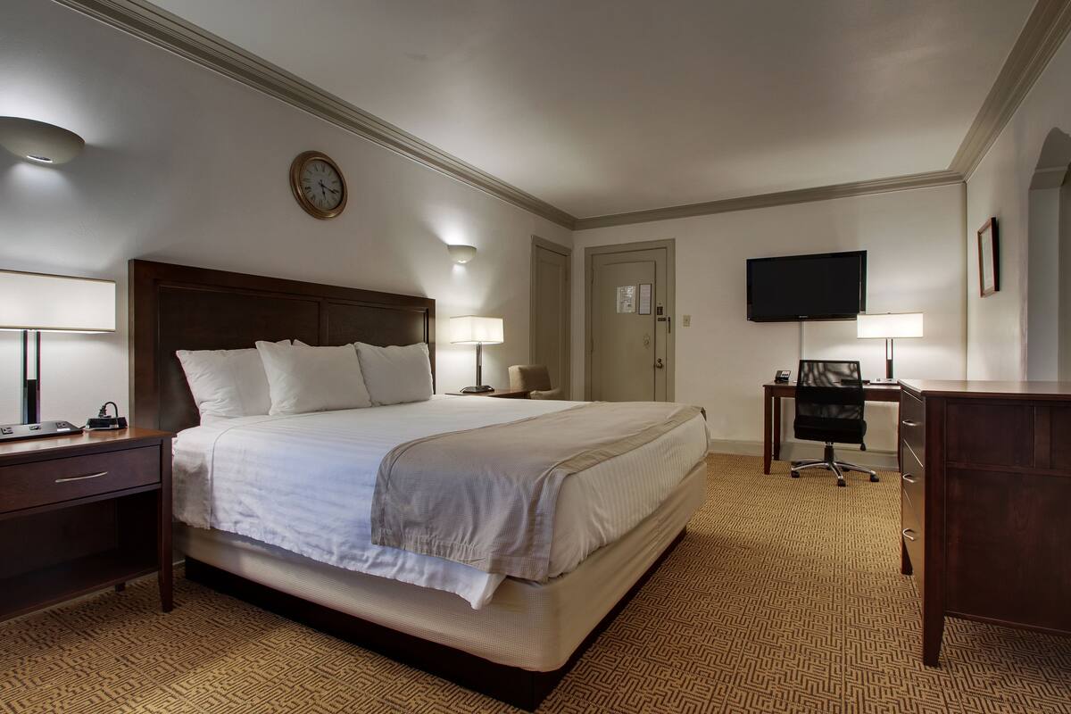 Ein Hotelzimmer mit einem großen Bett, Nachttischen, einem Schreibtisch mit Stuhl, einem Fernseher und einer Wanduhr.