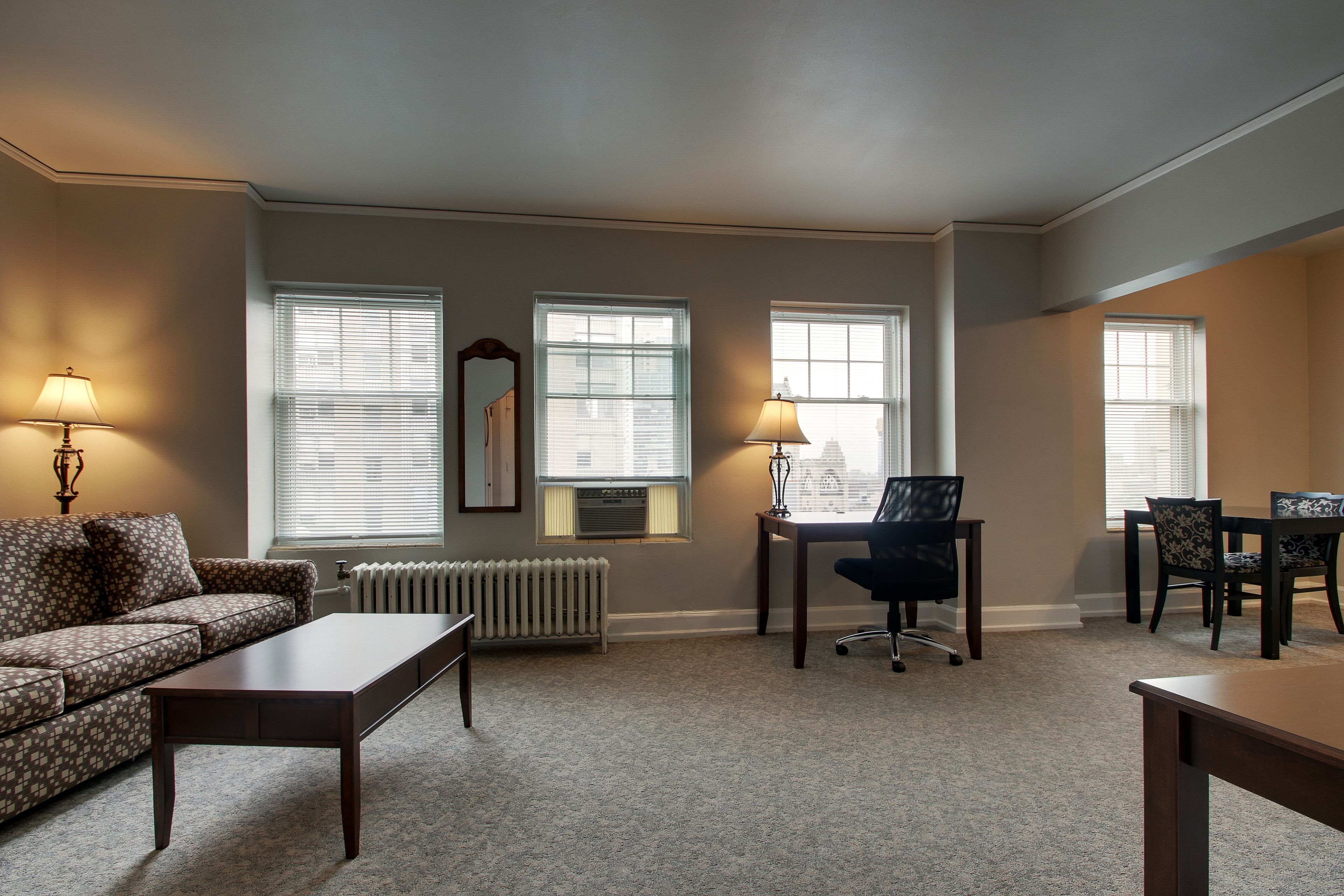 A living room with a sofa, coffee table, and dining table with chairs. There are three windows with blinds, a radiator, and a mirror on the wall.