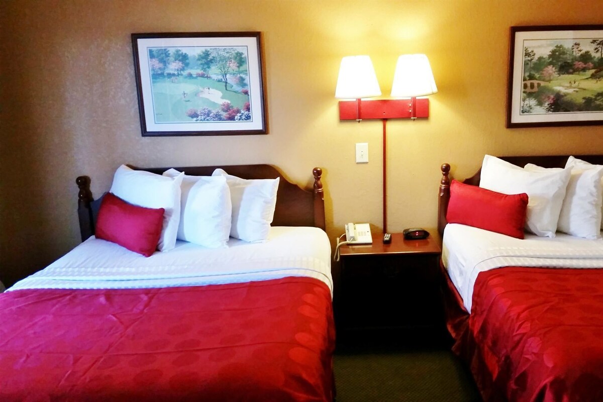 A hotel room with two beds, each with red bedding and white pillows, a nightstand with a lamp, and two framed landscape paintings on the walls.