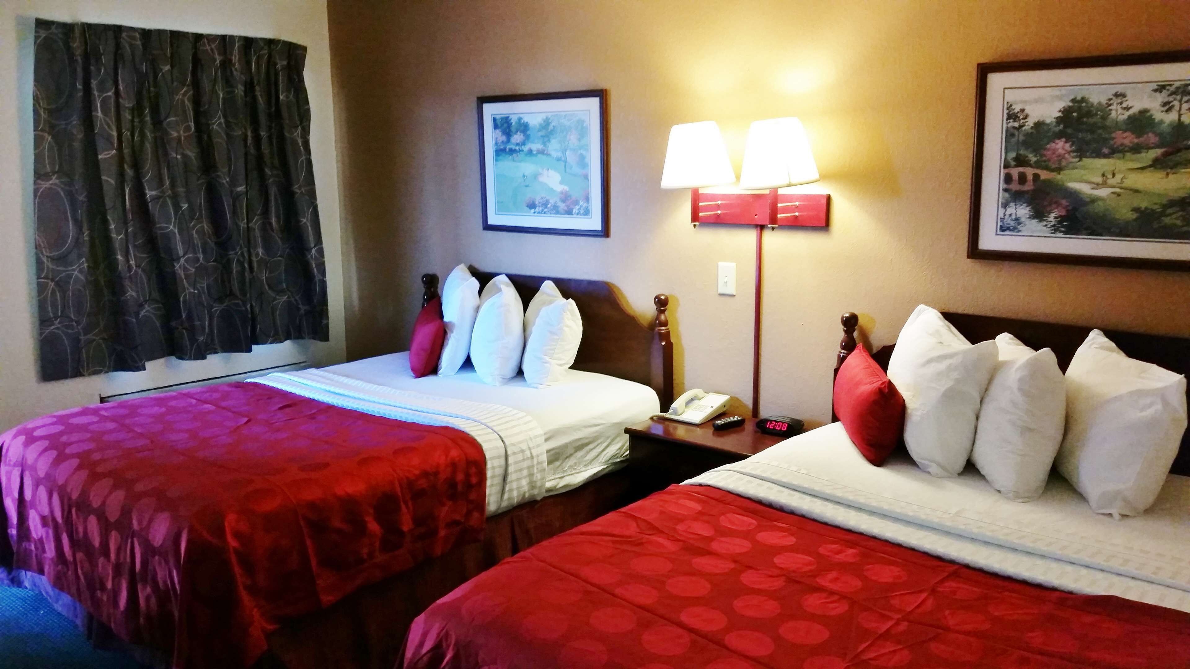 Une chambre d’hôtel avec deux lits, chacun étant agrémenté de draps rouges et d’oreillers blancs. On y trouve une table de chevet avec une lampe, un téléphone et une horloge. Un tableau est accroché au mur, au-dessus des lits.