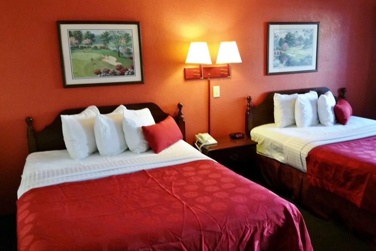 A hotel room with two beds, each with red bedding and white pillows, a nightstand with a lamp, and two framed pictures on the wall.