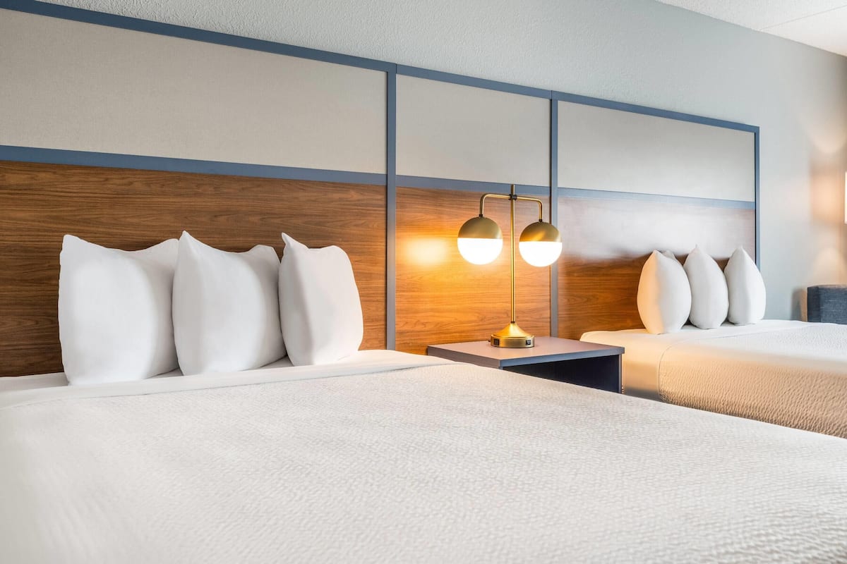 A hotel room with two beds, a wooden headboard, a nightstand with a lamp, and a wall panel design.