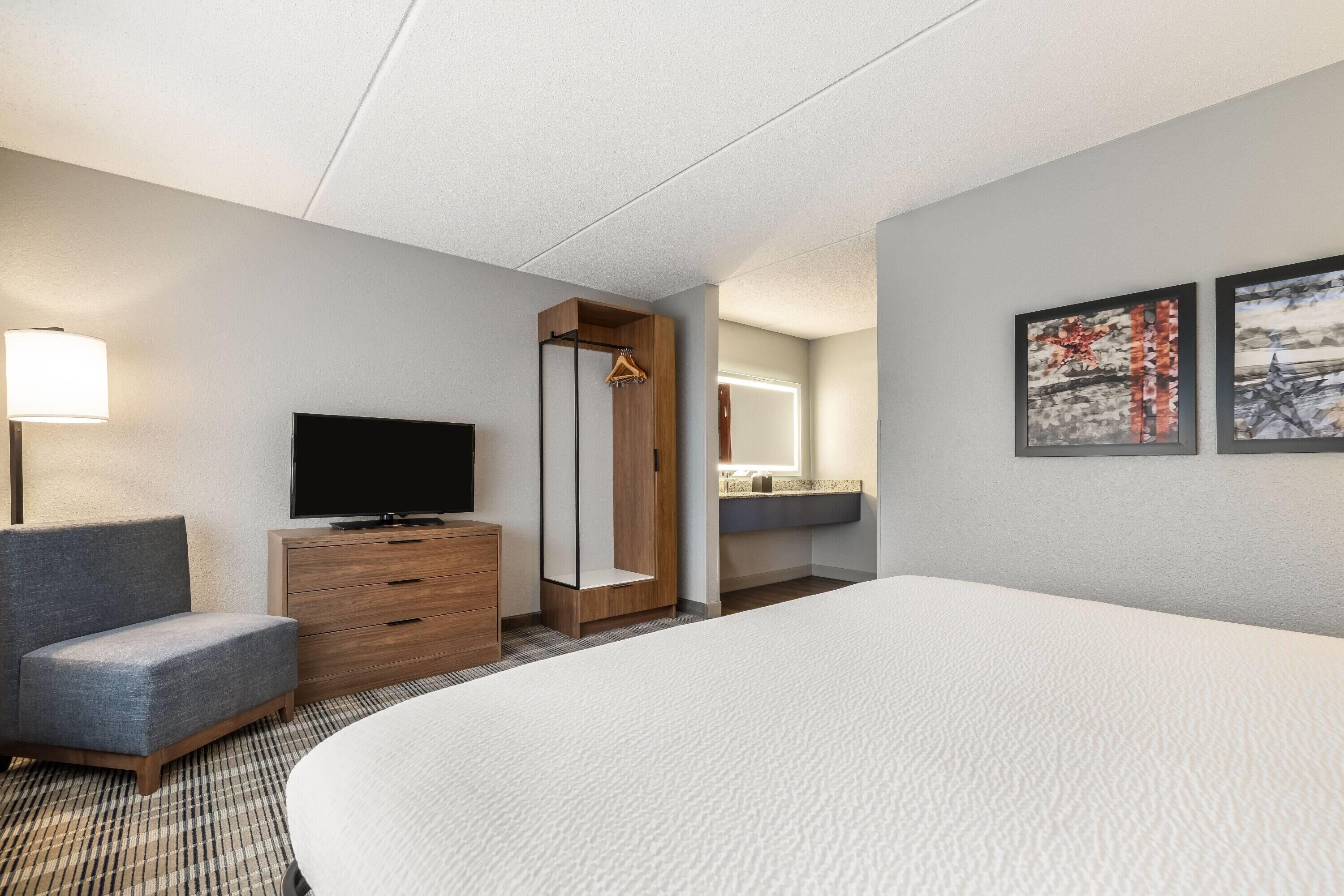 A hotel room with a bed, a TV, a wooden dresser, a gray armchair, a mirror, and two framed pictures on the wall.