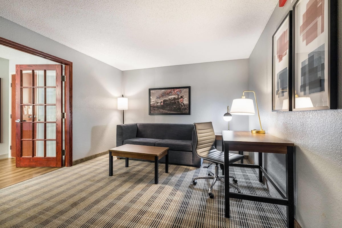 A hotel room with a sofa, a desk, two lamps, a framed picture on the wall, and a patterned carpet.