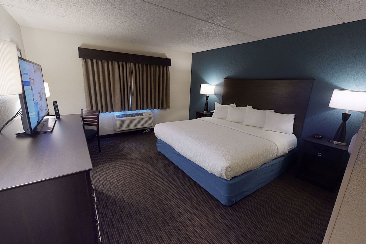 A hotel room with a bed, bedside tables, a desk with a computer, a chair, a window with curtains, and an air conditioning unit.