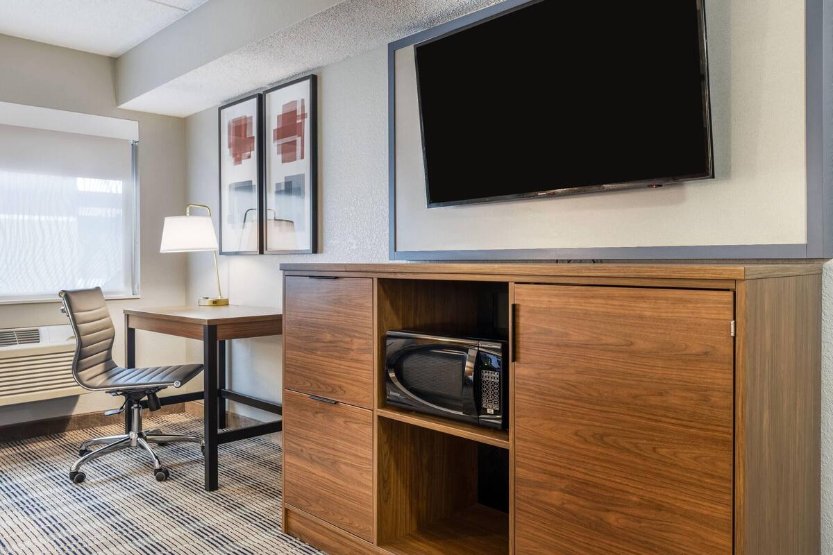 A hotel room with a wooden desk, a modern chair, a flat-screen TV, and a microwave in a wooden cabinet.