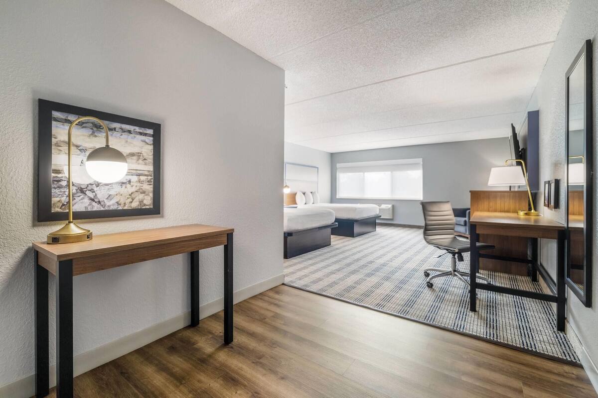 A hotel room with a wooden desk, a framed picture on the wall, a bed with a white headboard, a chair, and a desk lamp.