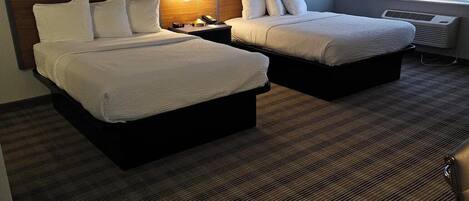 A hotel room with two beds, a headboard, bedside lamps, a window with blinds, and a carpeted floor.