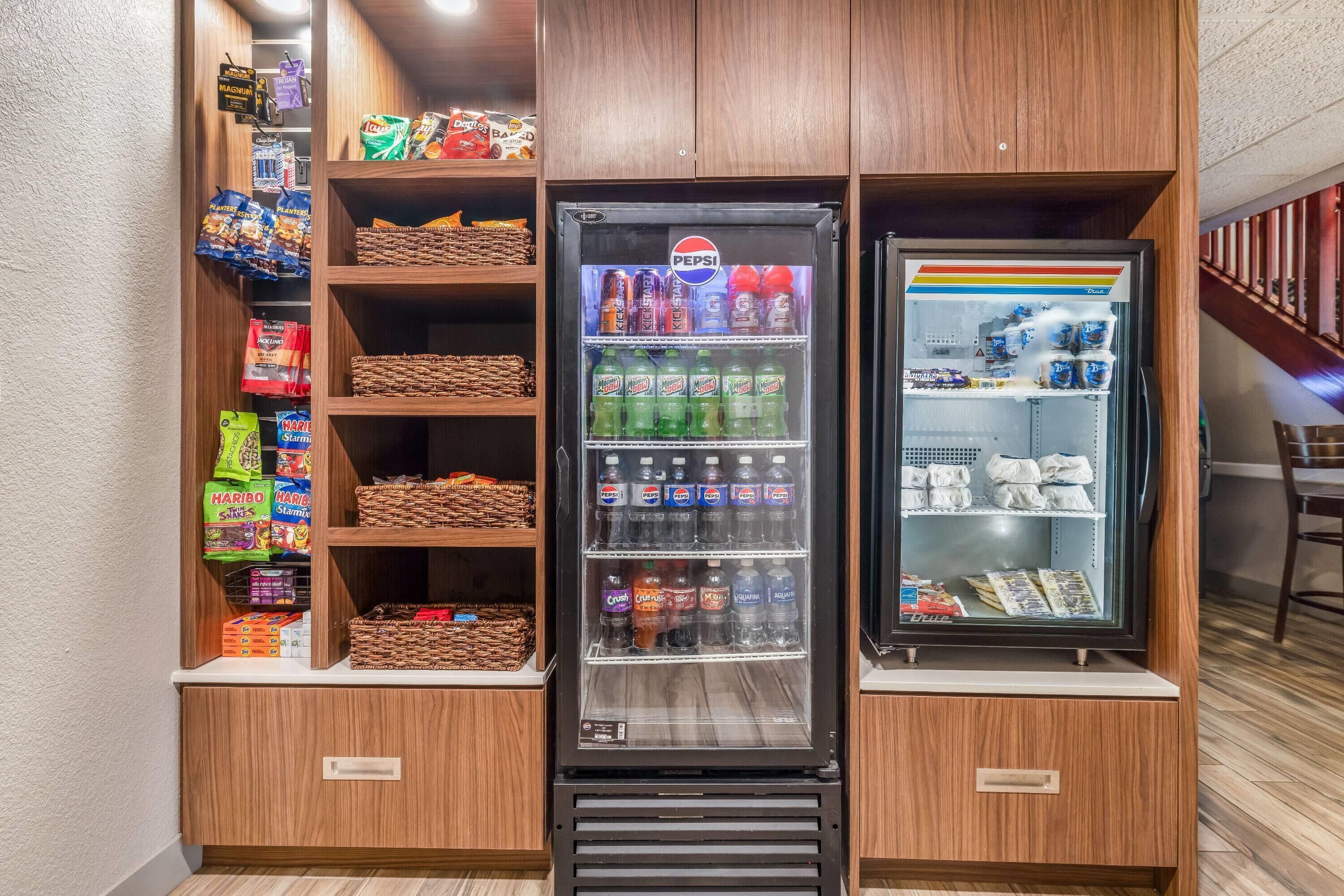 Zona de máquinas expendedoras con un refrigerador de Pepsi y una máquina de Coca-Cola, rodeada de estantes llenos de aperitivos y bebidas.