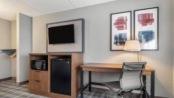 Habitación de hotel con televisión de pantalla plana, escritorio con silla, microondas y refrigerador. Se observan pinturas abstractas en la pared y una lámpara sobre el escritorio.