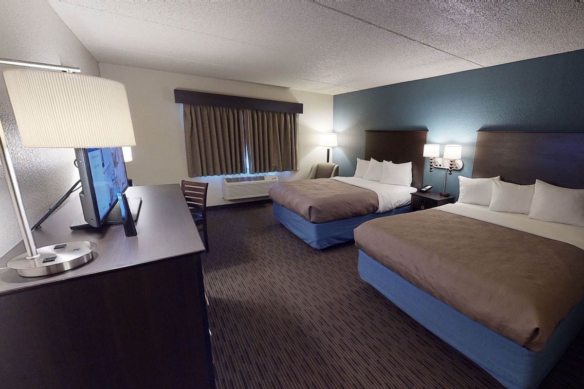 A hotel room with two beds, a desk, a chair, a lamp, and a window with curtains.