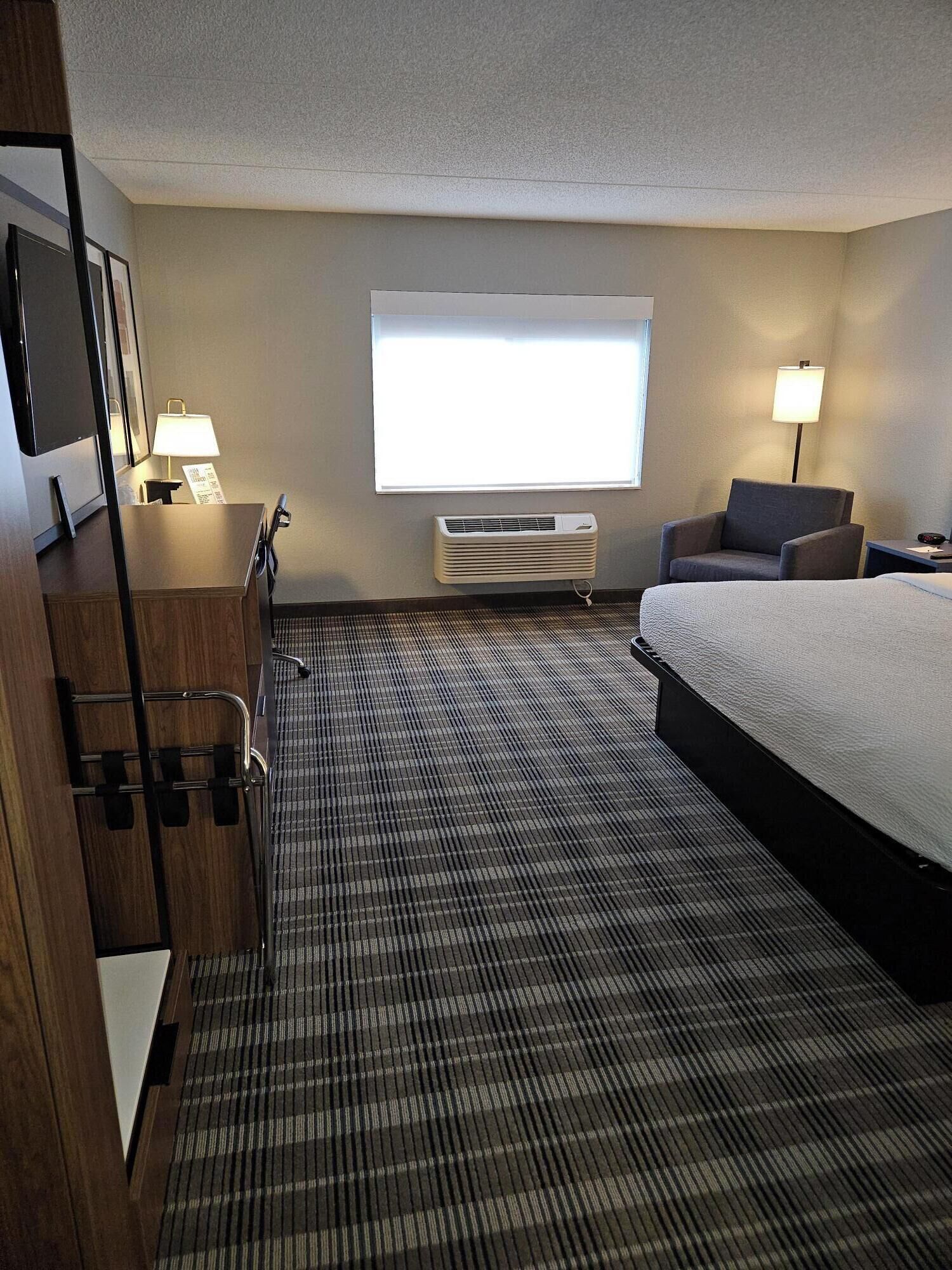 A hotel room with a bed, a desk, a chair, a TV, and a window with blinds.