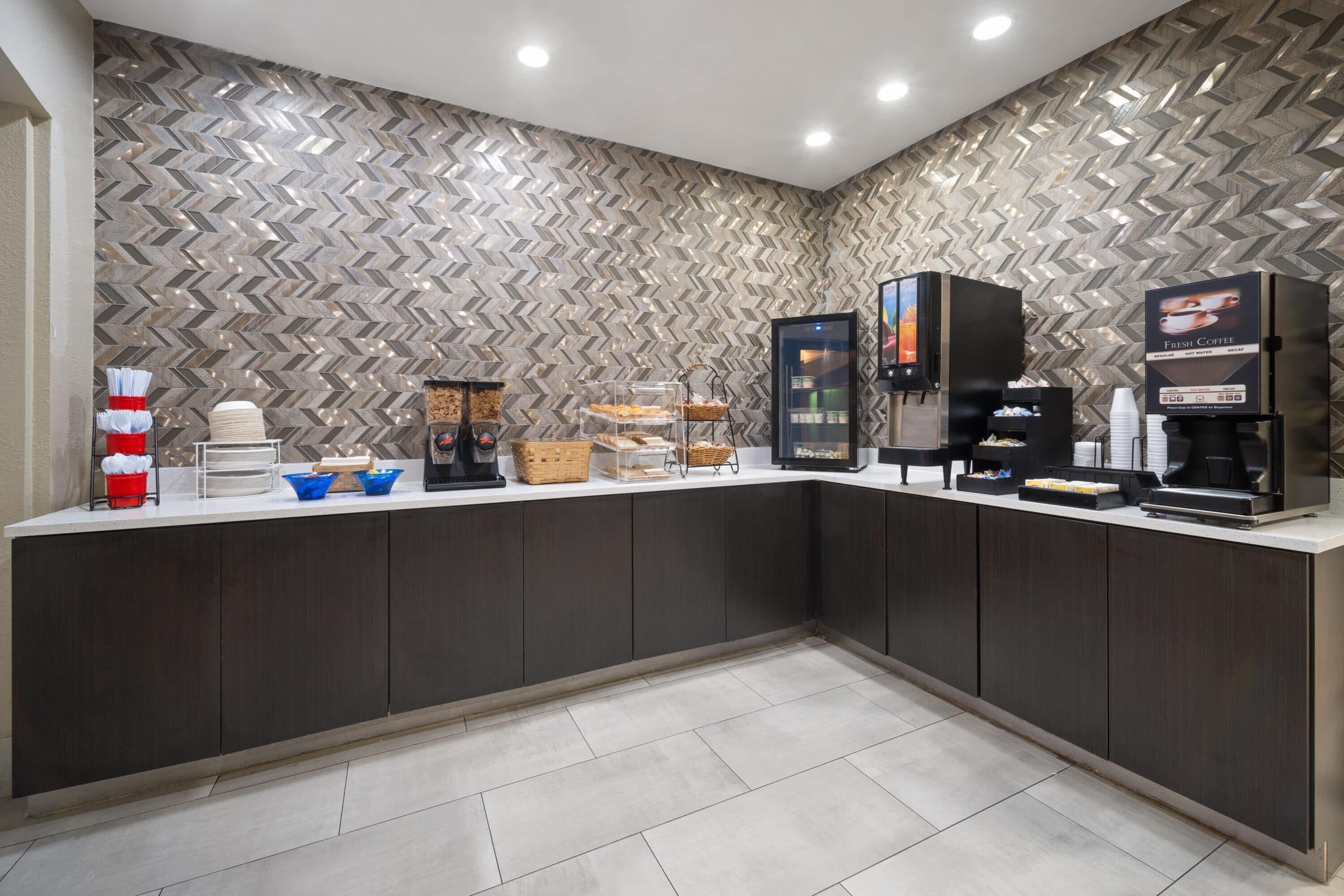 A modern hotel reception area with a herringbone-patterned wall, a self-service coffee station, and a counter with various amenities.