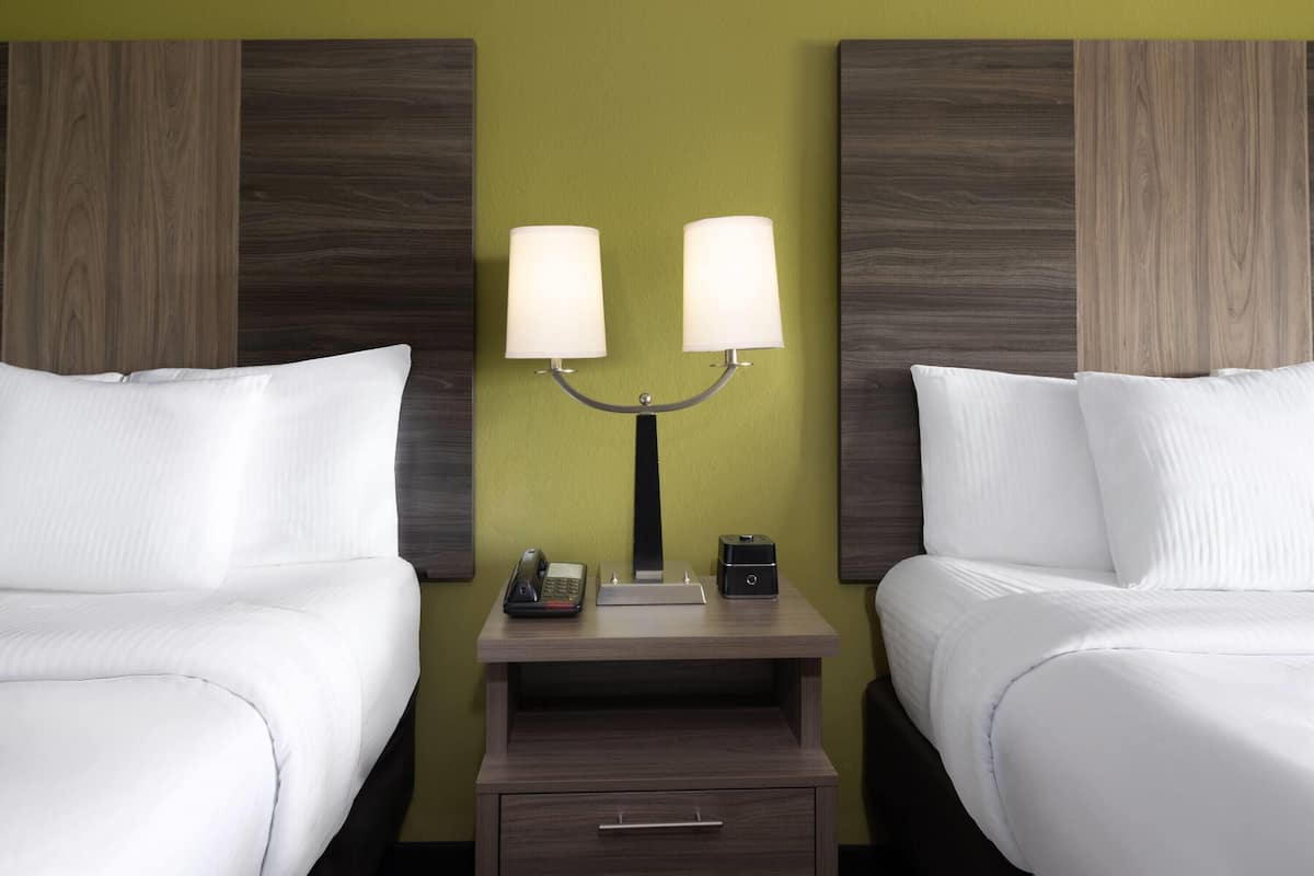 A hotel room with two beds, a nightstand with a lamp, a telephone, and a small safe.