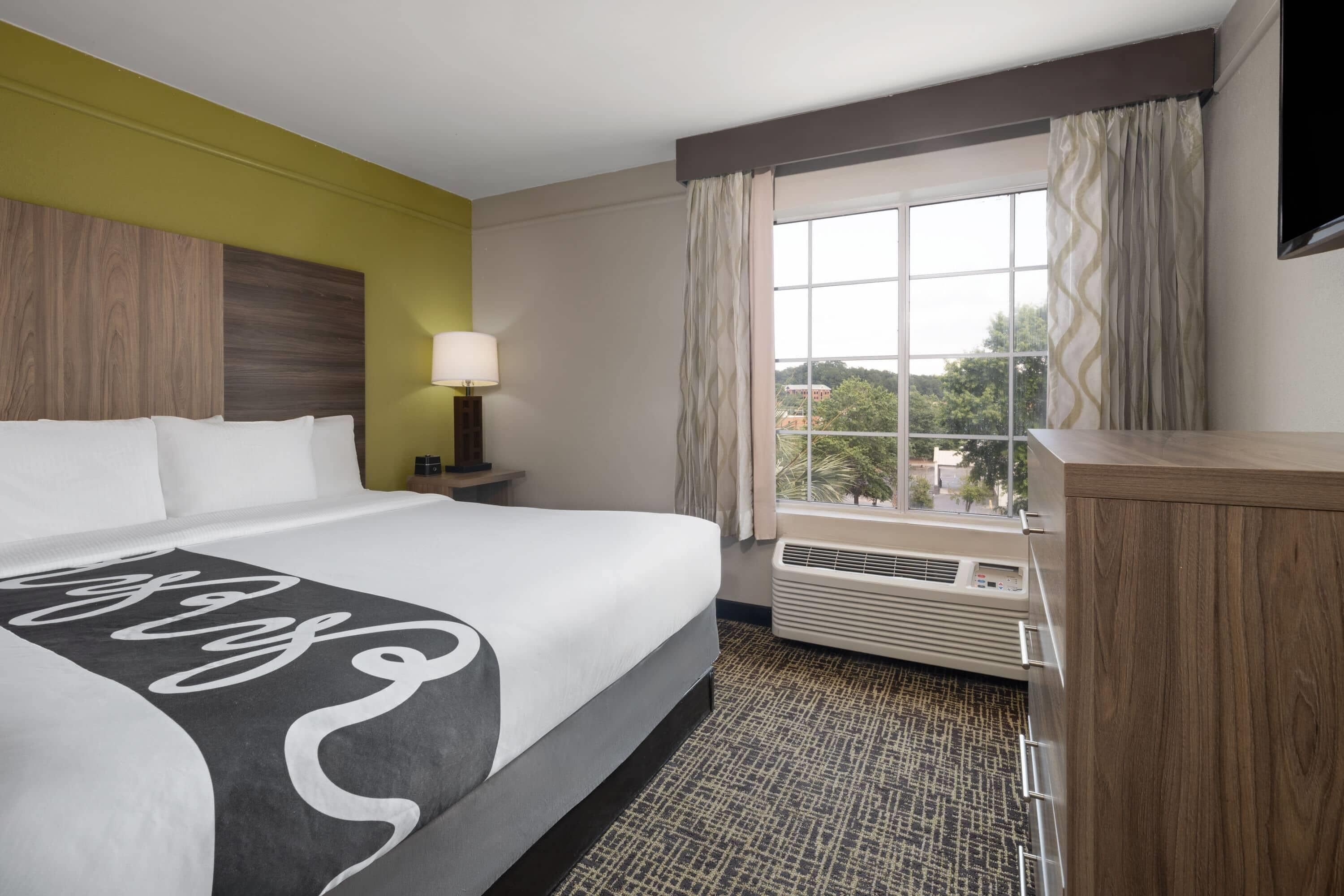Una habitación de hotel con una cama grande, un escritorio, una lámpara, una ventana con cortinas y un equipo de aire acondicionado.