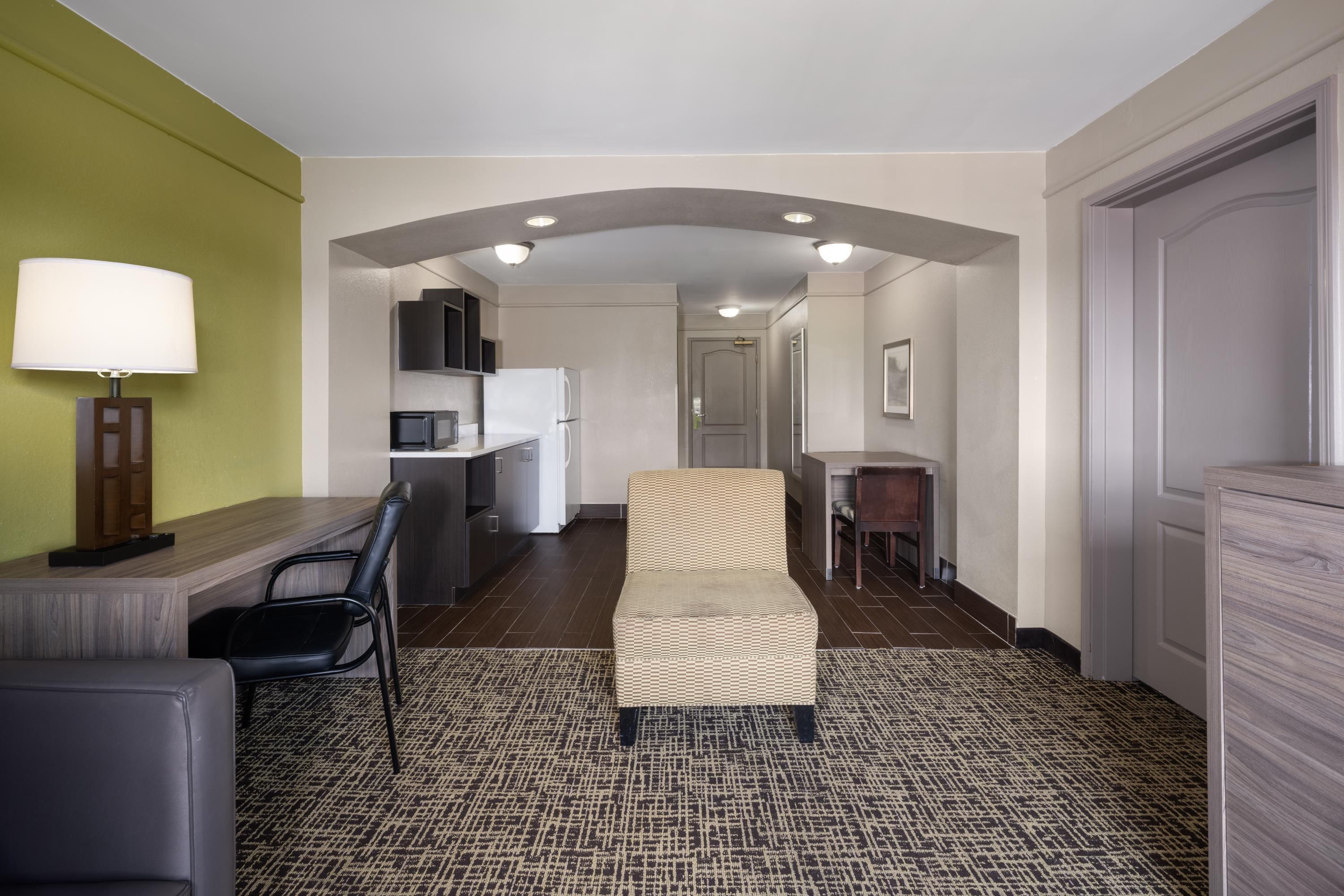 A hotel room with a kitchenette, a seating area with a sofa and chairs, a patterned carpet, and a lamp.