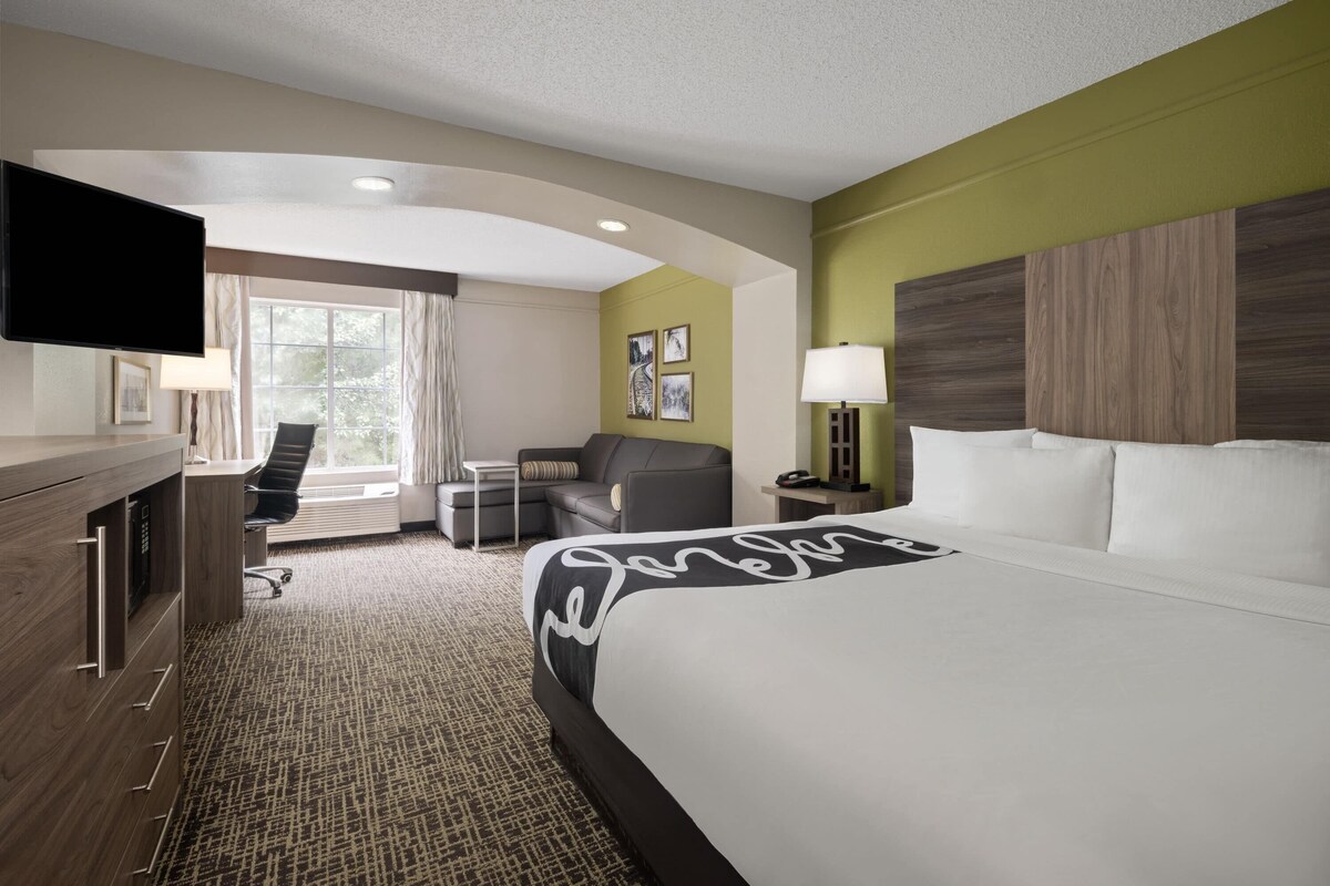 A hotel room with a bed, desk, chair, TV, and artwork on the wall.