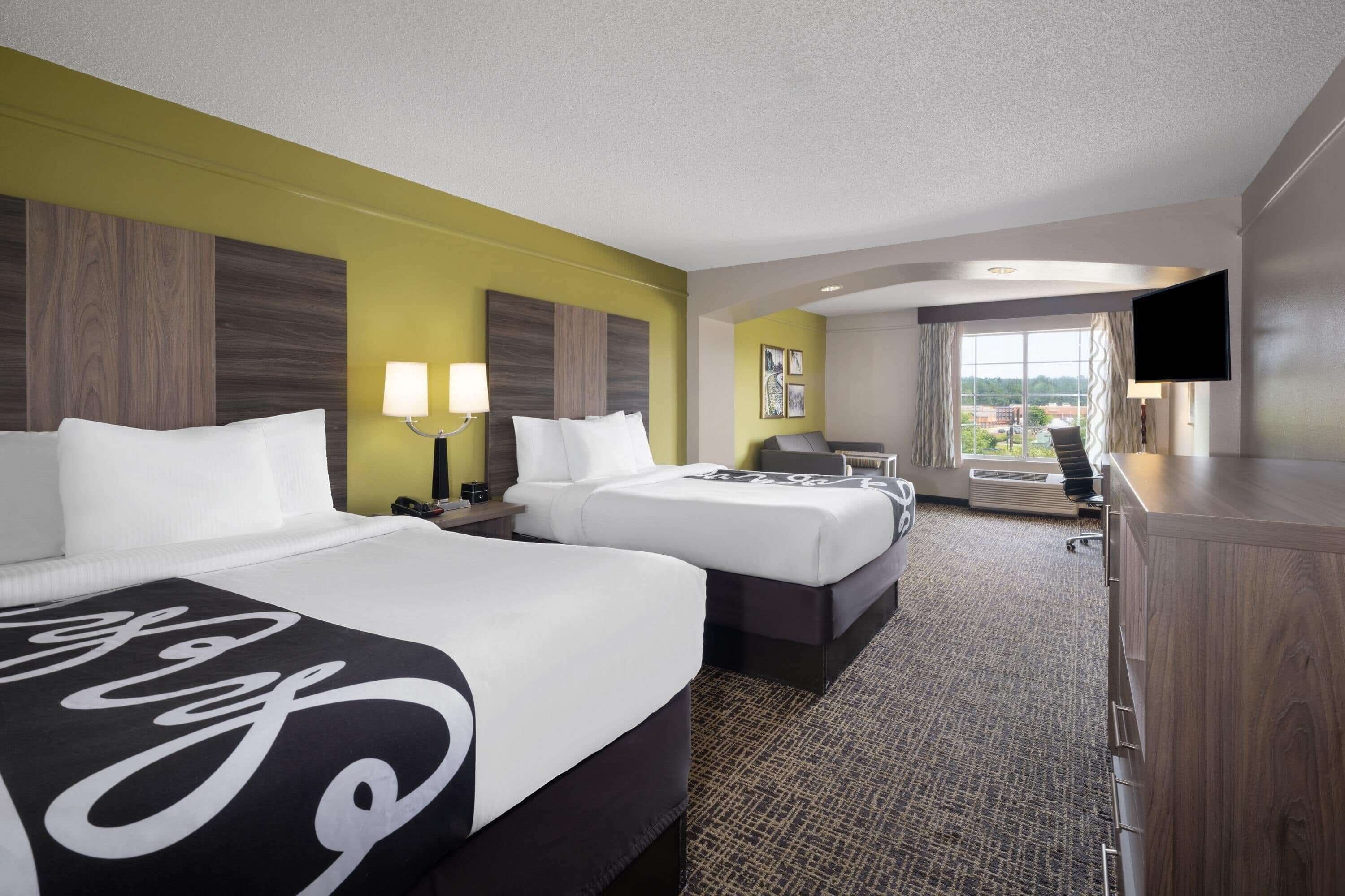 A hotel room with two beds, a desk, a TV, and a large window.