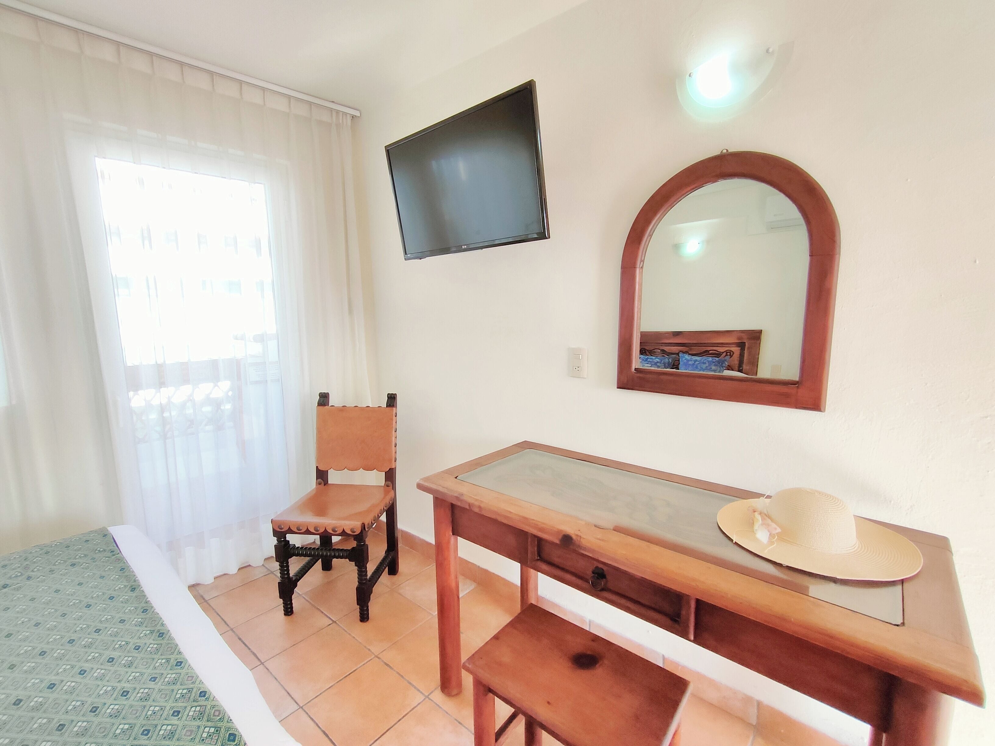 A room with a bed, a wooden desk, a chair, a television mounted on the wall, and a mirror.