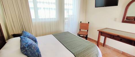 Quarto de hotel com cama, escrivaninha, cadeira, televisão e janela com cortinas.