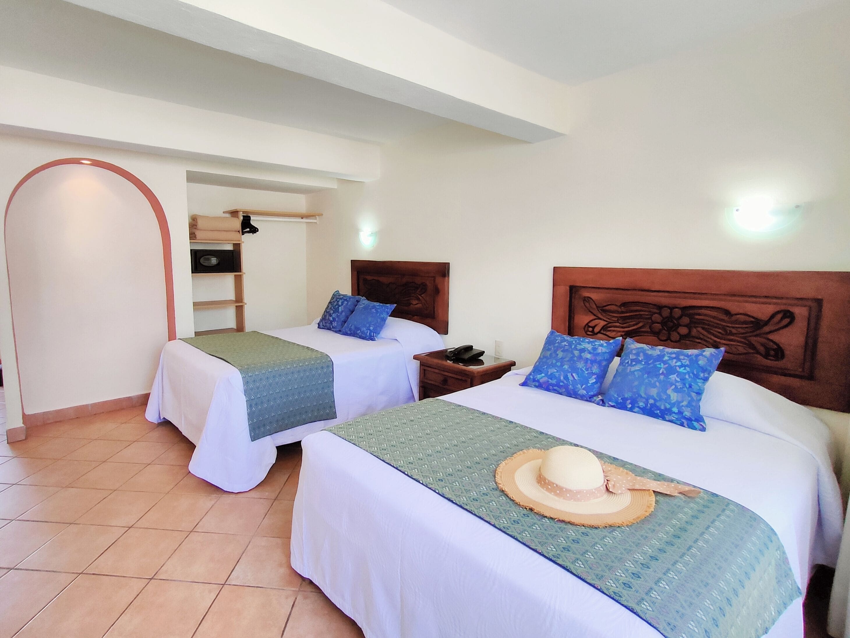 Habitación de hotel con dos camas, cada una con cabecero de madera, una mesita de noche y una mesilla auxiliar. Se observa una pequeña estantería con algunos objetos y un televisor montado en la pared.
