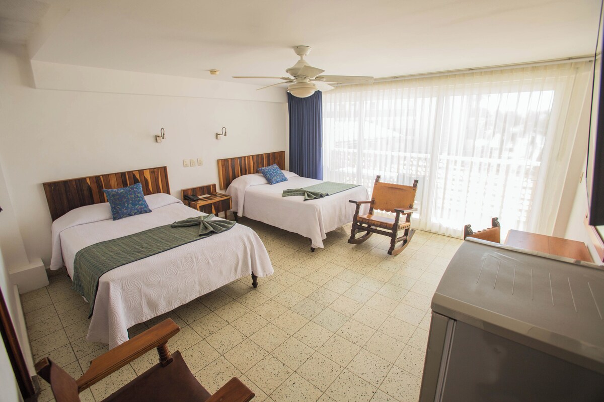 Une chambre d’hôtel avec deux lits, un ventilateur de plafond, un fauteuil à bascule en bois et une kitchenette.