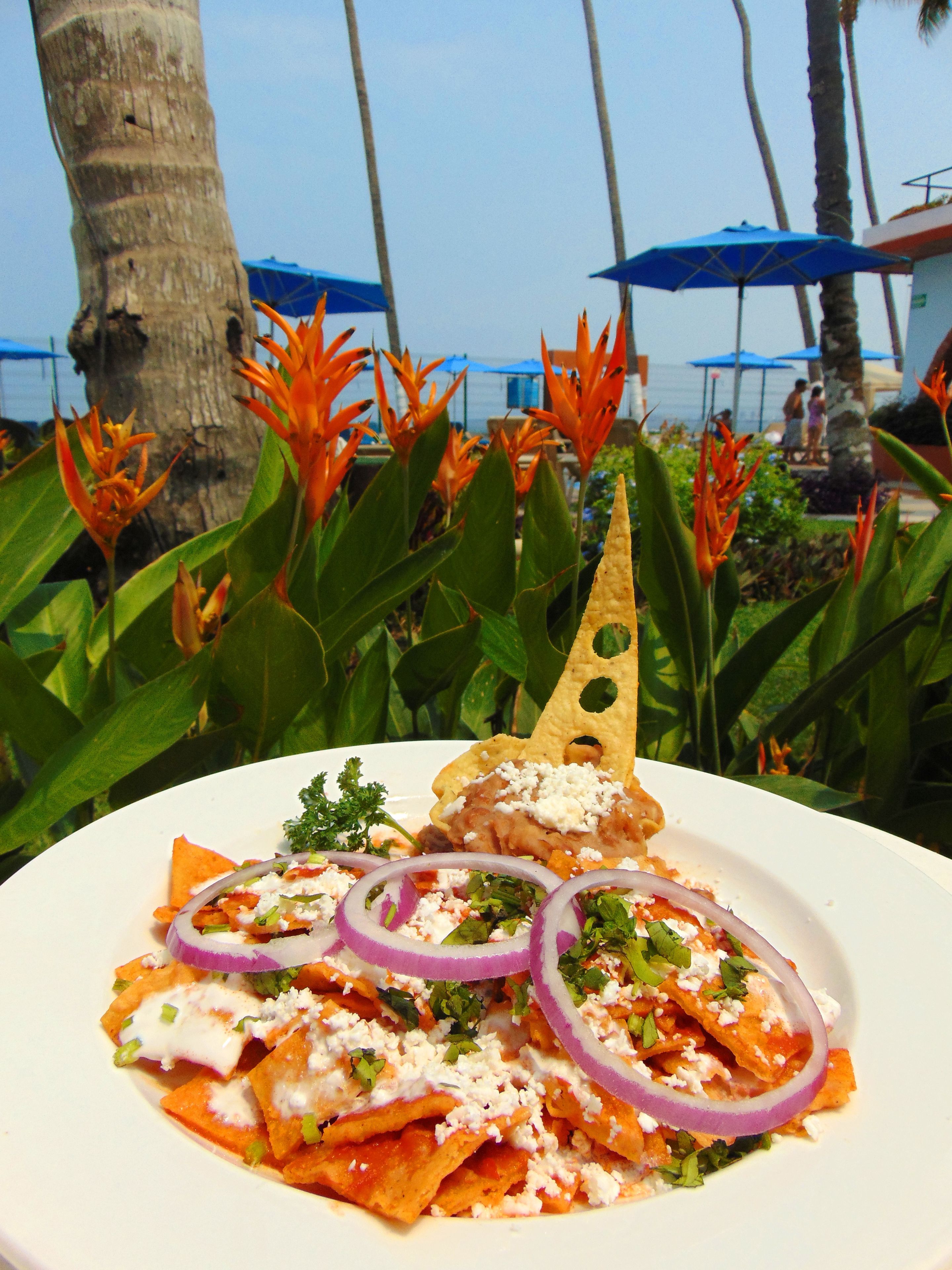 A plate of nachos with toppings, set against a backdrop of tropical plants and a beach view.