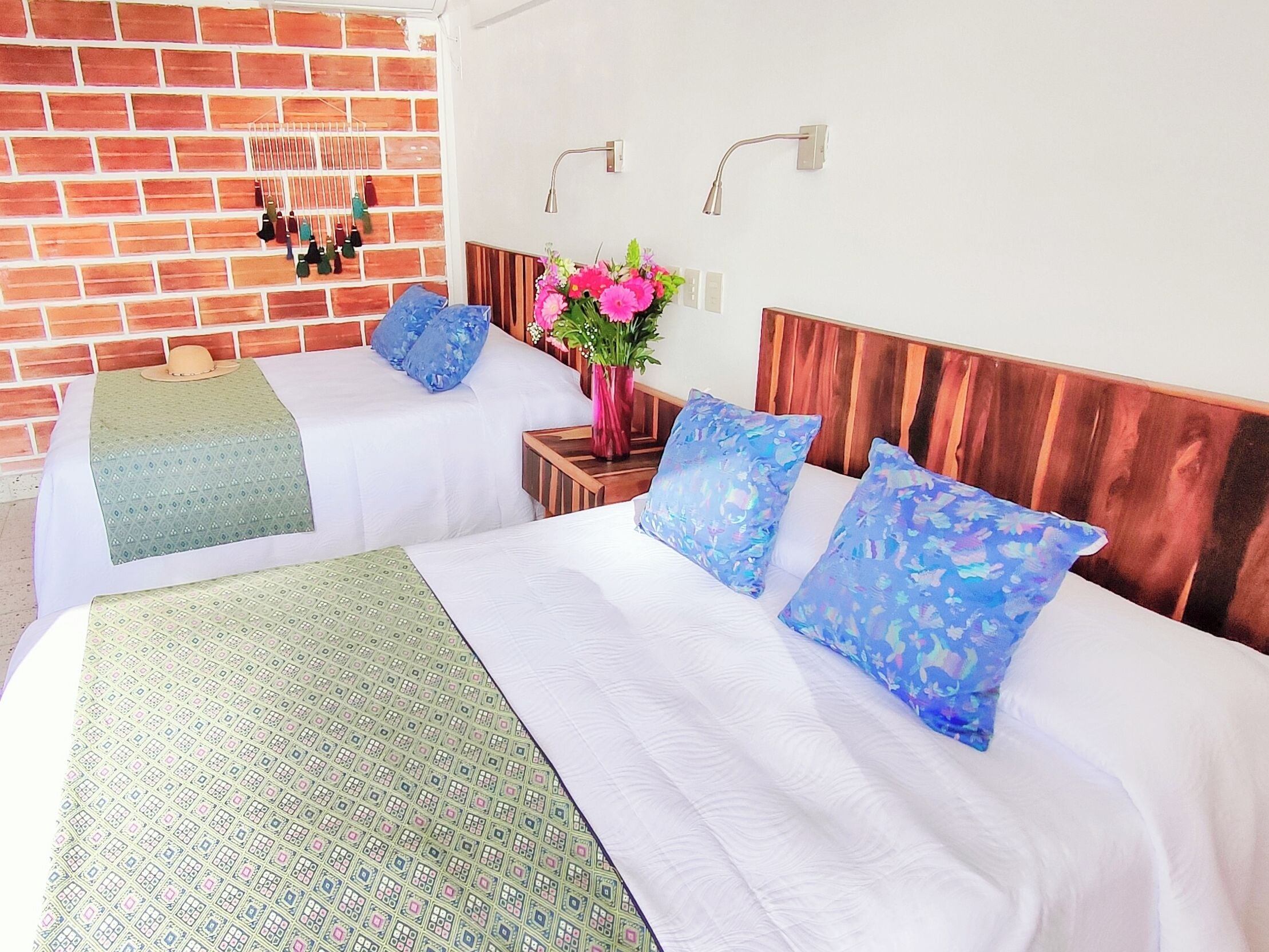 A hotel room with two beds, a brick wall, a wooden headboard, a nightstand with a vase of flowers, and a wall-mounted light fixture.