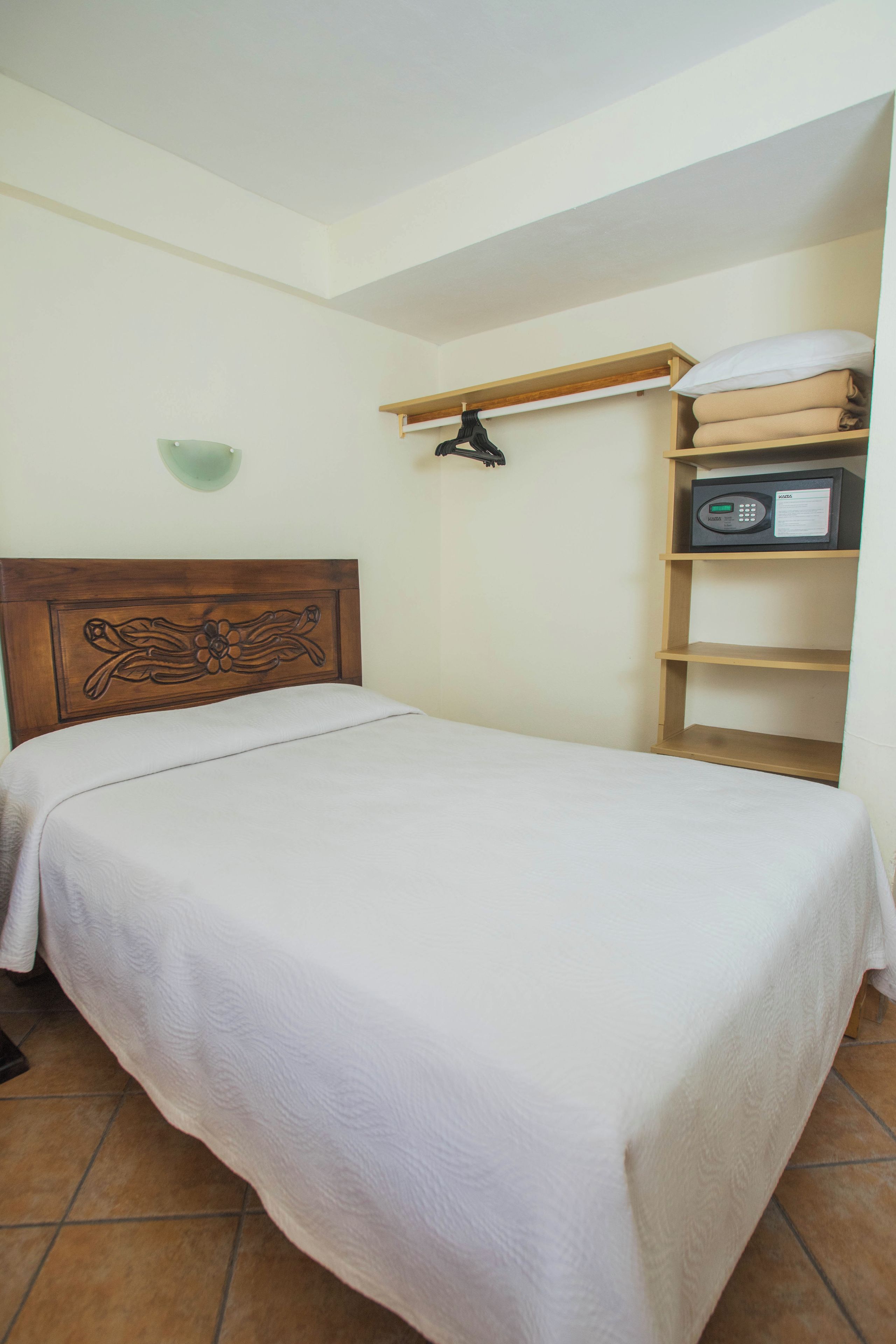 Een slaapkamer met een houten hoofdbord, een bed met een witte spreideken, een plank met opgevouwen handdoeken, en een magnetron aan de muur.