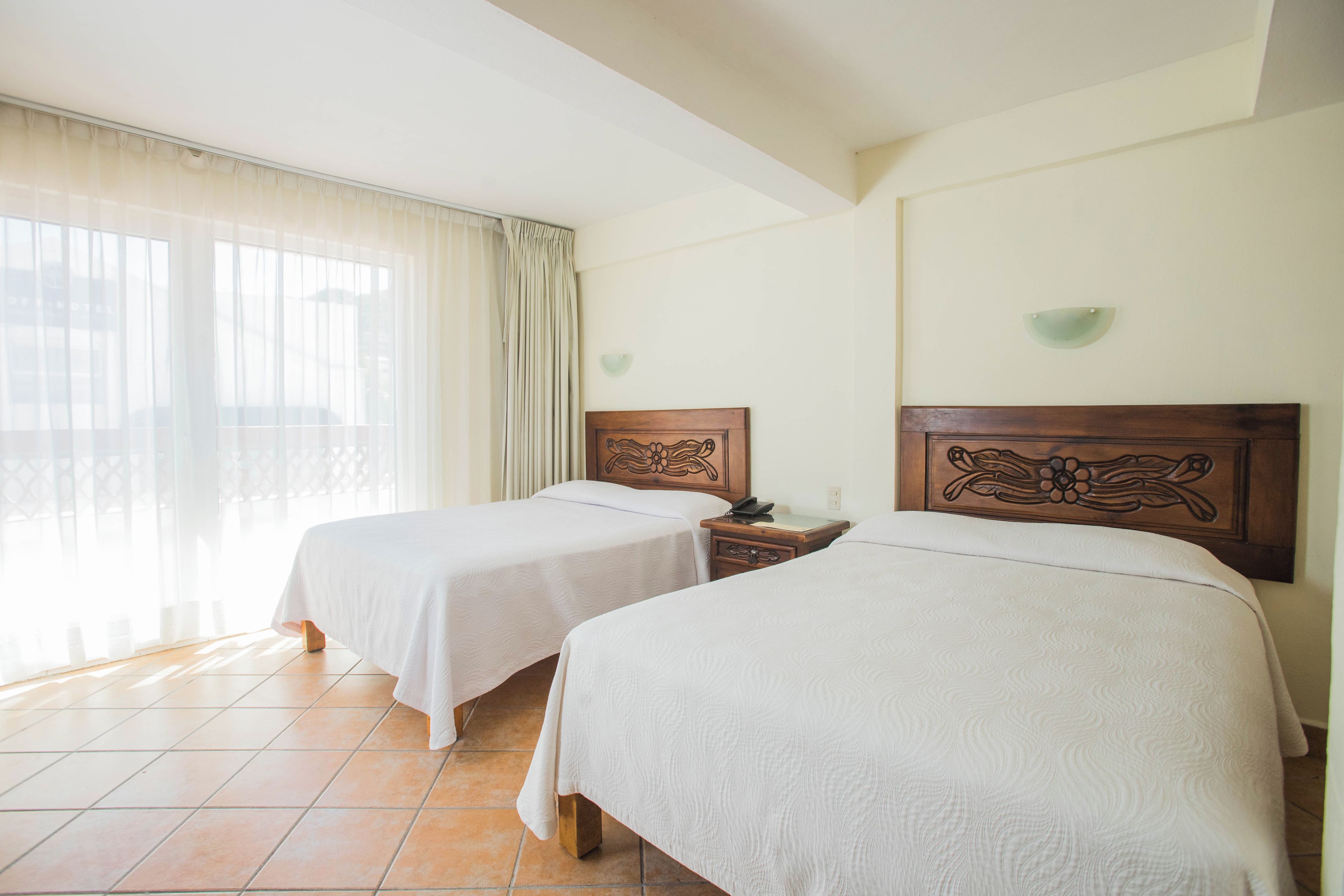 A hotel room with two beds, a nightstand, a lamp, and a window with curtains.