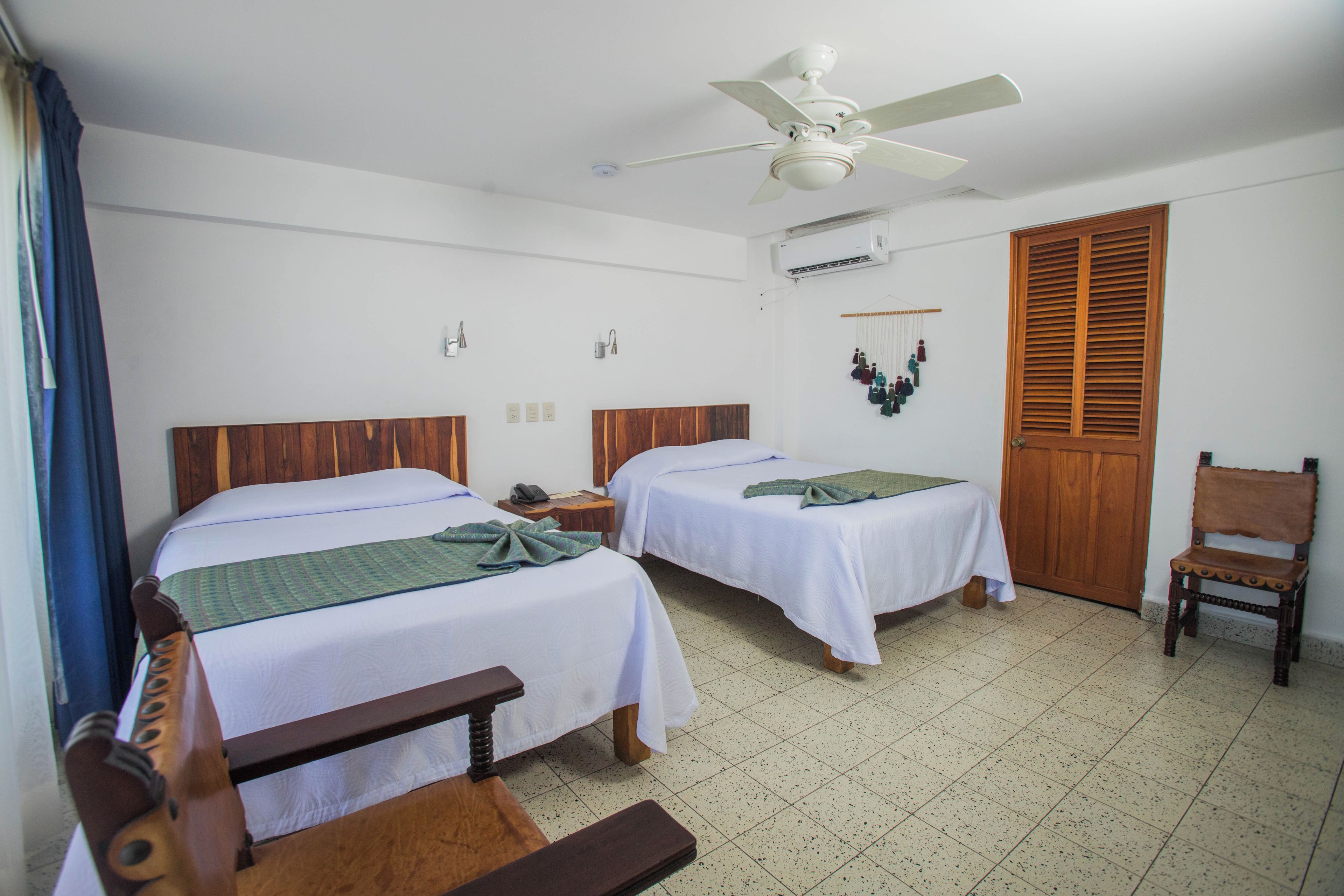 Habitación de hotel con dos camas, ventilador de techo, aire acondicionado, puerta de madera y una silla.